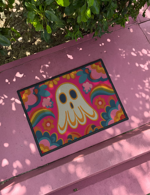 A rectangular polyester doormat with a vibrant, psychedelic design on a hot pink background. In the center is a cute, cartoonish ghost in off-white with yellow and orange outlines; it has large, dark, expressive eyes. The ghost is surrounded by a maximalist pattern of 70s-style floral motifs and puffy clouds in shades of teal, blue, orange, and light pink. Two small, colorful rainbows arc over the ghost's shoulders. The doormat has a durable, textured surface and is framed by a sleek black trim.